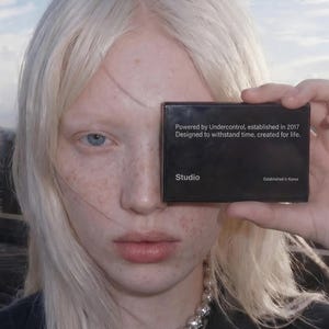 May include: A person with pale skin and blonde hair holds a black card in front of their face. The card reads "Powered by Undercontrol, established in 2017. Designed to withstand time, created for life. Studio." A silver necklace is visible.