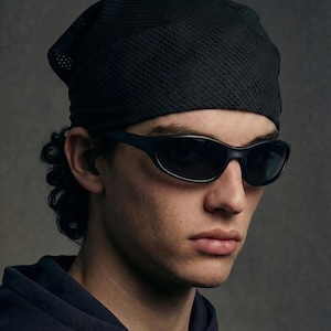 May include: Black sunglasses with a sleek, curved design. The person in the image is wearing a black mesh bandana and a dark blue hoodie. The background is a solid, neutral gray.