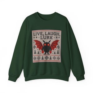 May include: Dark green sweatshirt with a festive design. The design features a black creature with red wings and glowing eyes, with the text "LIVE, LAUGH, LURK" above it. The design is framed by a pattern of snowflakes and trees.
