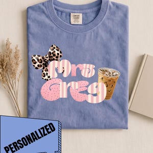 Puede incluir: Una camiseta de manga corta color azul bígaro con una etiqueta Comfort Colors en el cuello. El gráfico presenta un lazo con estampado de leopardo, el texto "Mrs Greg" en letras a rayas rosas y blancas, y un vaso con café helado.