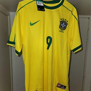 Może przedstawiać: Żółta koszulka piłkarska Brazylii z zielonymi akcentami na rękawach i kołnierzu. Numer 9 jest wydrukowany na czarno z przodu. Widoczne jest logo Nike i herb reprezentacji Brazylii. Koszulka wisi na czarnym wieszaku.