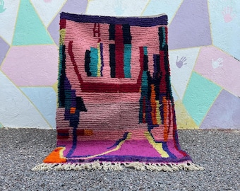 May include: A colorful, hand-woven rug with a geometric design. The rug features a pink and purple base with accents of red, teal, orange, and yellow. The edges are trimmed with a fringe. The rug is displayed against a multi-colored wall.