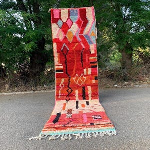 Op de afbeelding: Een levendig, rechthoekig tapijt met een geometrisch ontwerp. Het tapijt heeft een rode achtergrond met patronen in roze, oranje, zwart en wit. Het bovenste gedeelte heeft ruitvormen in verschillende kleuren. De onderste rand heeft een franje met blauwe en witte accenten.