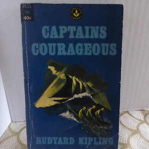 May include: A vintage paperback book titled "Captains Courageous" by Rudyard Kipling. The cover is a deep blue with white and yellow illustrations of a ship at sea. The book is from the Laurel-Leaf Library.