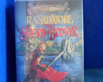 May include: A paperback book cover with a fantasy illustration of a man in red armor holding a sword. The title of the book is "The Sword of Bedwyr" by R.A. Salvatore. The book is part of the "The Crimson Shadow" series.