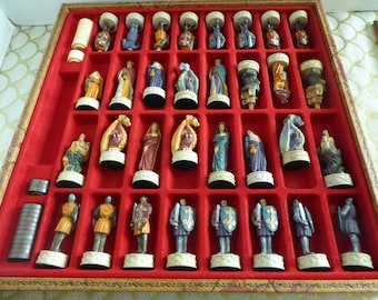 May include: A set of chess pieces in a wooden box. The pieces are made of painted resin and feature a medieval theme. The pieces are arranged in their starting positions on a red felt board.