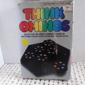 May include: A boxed Think Ominos solitaire game. The box is gray with the game title in rainbow colors. The game features a black, six-sided domino teaser with colored dots. The text on the box reads, "Solve this six-sided domino teaser by matching color dots to facing color dots."