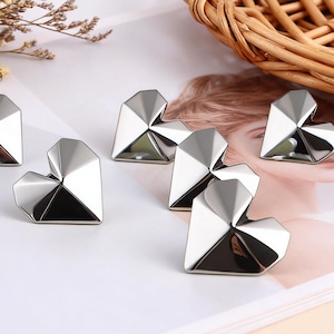 May include: A collection of silver heart-shaped paper clips arranged on a white surface. The clips have a geometric design with multiple facets, reflecting light. Dried flowers and a wicker basket are in the background.