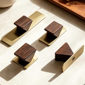 May include: Four cabinet knobs and pulls featuring a combination of dark brown wood blocks and brushed gold metal rectangular backplates. The hardware pieces vary in length, with some featuring elongated plates and others square bases.