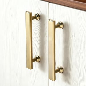 May include: Two vertical, rectangular brass cabinet pulls with a polished finish, mounted on white wood-grain cabinet doors. Each handle features a flat, elongated bar design secured by two circular posts, providing a clean and functional aesthetic.