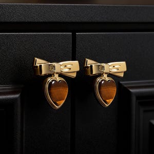 May include: Two gold cabinet knobs with a bow design and heart-shaped tiger's eye stone centres. The knobs are mounted on a black cabinet door. The hardware has a polished finish.