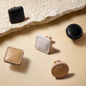 Pode incluir: Uma variedade de puxadores de couro para armários em várias formas e cores. Os puxadores incluem designs quadrados e redondos em preto, bege e creme, com costuras brancas e ferragens douradas. Estes puxadores decorativos são ideais para móveis.