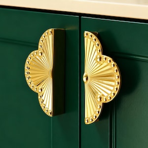 May include: Close-up of two gold-colored cabinet handles. Each handle has a fan-like design with a beaded border and radial lines. The handles are mounted on a dark green cabinet door.
