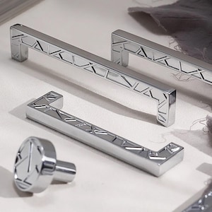 Può includere: Un set di ferramenta per mobili cromata lucida, tra cui due maniglie rettangolari, una maniglia a forma di L e un pomello rotondo. L'hardware presenta un design geometrico con intagli angolari, che riflettono la luce. Gli articoli sono disposti su una superficie chiara.