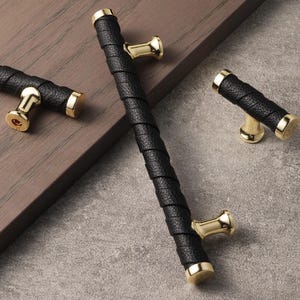 May include: Black leather-wrapped cabinet pulls with gold-toned accents. The hardware features a textured leather grip and cylindrical gold end caps. The pulls have a modern design and are suitable for various furniture pieces.