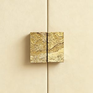 May include: A close-up shot of a cabinet door featuring two square brass handles. The handles have a textured, organic design and a warm gold finish. The cabinet door is a light cream colour, with the handles positioned centrally.