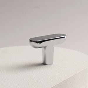 May include: A shiny, chrome-finished cabinet knob with a T-shaped design. The knob features a rectangular top and a cylindrical base, reflecting light. This hardware is suitable for drawers and cabinets.