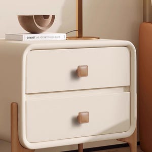 Może przedstawiać: Kremowy stolik nocny z dwiema szufladami, z kwadratowymi, brązowymi sk&oacute;rzanymi uchwytami. Stolik nocny ma zaokrąglone krawędzie i stoi na jasnych drewnianych nogach. Na g&oacute;rze książka i dekoracyjna miska.