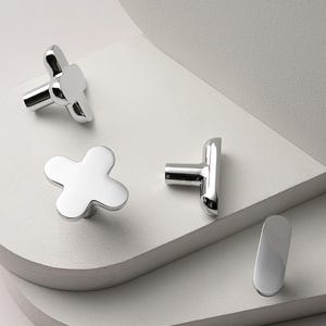 May include: A collection of shiny, chrome-finished cabinet hardware. The set includes three flower-shaped knobs and one elongated, curved handle. The hardware is displayed on a white, layered surface, showcasing the reflective quality of the metal.