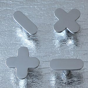 May include: Four silver-colored cabinet knobs in the shape of mathematical symbols: a plus sign, a minus sign, a multiplication sign, and a division sign. The knobs are set against a textured, silver background.