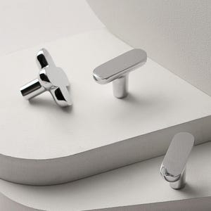 May include: A collection of three modern, silver-toned cabinet knobs. One knob has a four-petal design, while the other two have a sleek, elongated shape. The knobs are displayed on a white, layered surface.