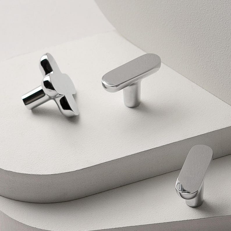 May include: A collection of three modern, silver-toned cabinet knobs. One knob has a four-petal design, while the other two have a sleek, elongated shape. The knobs are displayed on a white, layered surface.