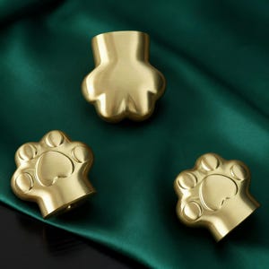 May include: Three brass paw-shaped cabinet knobs. The knobs are gold-colored and have a smooth, polished finish. The background is a dark green fabric.