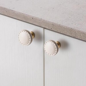 Może przedstawiać: Dwa okrągłe uchwyty do szafek z jasnobeżową powierzchnią przypominającą sk&oacute;rę i białymi przeszyciami. Gałki mają podstawę w kolorze mosiądzu i są zamontowane na białych drzwiach szafki. Szafka znajduje się obok szarego blatu.