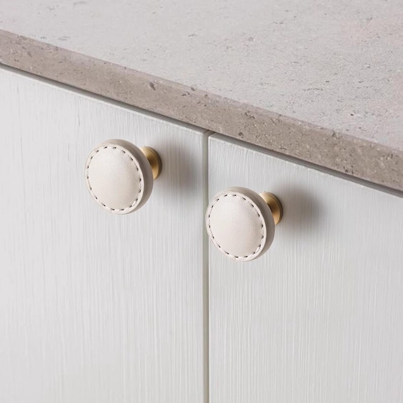 Może przedstawiać: Dwa okrągłe uchwyty do szafek z jasnobeżową powierzchnią przypominającą sk&oacute;rę i białymi przeszyciami. Gałki mają podstawę w kolorze mosiądzu i są zamontowane na białych drzwiach szafki. Szafka znajduje się obok szarego blatu.