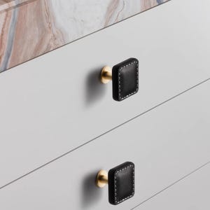 Może przedstawiać: Zbliżenie dw&oacute;ch czarnych, kwadratowych gałek do szafek z białymi przeszyciami i podstawami w kolorze złotym. Gałki są zamontowane na jasnoszarej szafce z marmurowym blatem w tle. Gałki są r&oacute;wnomiernie rozmieszczone.