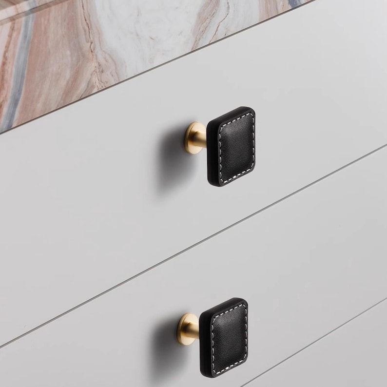 Może przedstawiać: Zbliżenie dw&oacute;ch czarnych, kwadratowych gałek do szafek z białymi przeszyciami i podstawami w kolorze złotym. Gałki są zamontowane na jasnoszarej szafce z marmurowym blatem w tle. Gałki są r&oacute;wnomiernie rozmieszczone.