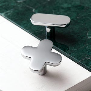 May include: Two polished chrome cabinet knobs. One is a four-leaf clover shape, and the other is a rectangular bar shape. The knobs are set against a white and green marble surface.