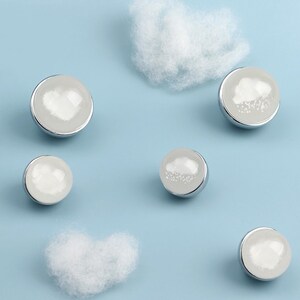 May include: A collection of silver-toned, round buttons with a white, cloud-like design on a light blue background. Fluffy white cotton clouds are scattered around the buttons, creating a whimsical, weather-themed aesthetic.