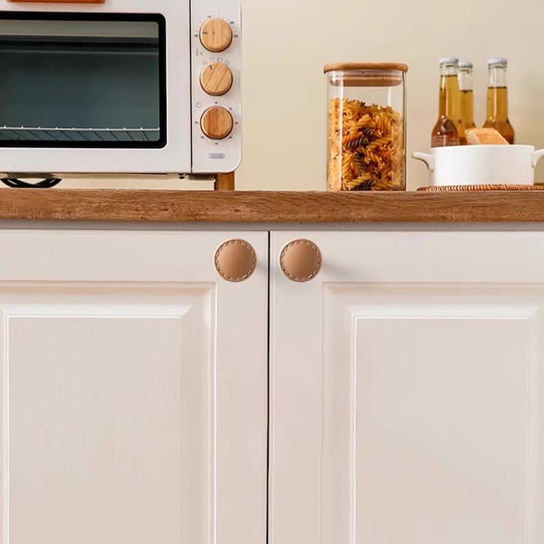 Może przedstawiać: Scena kuchenna z białym piekarnikiem z drewnianymi pokrętłami, szklanym słoikiem wypełnionym makaronem i dwoma jasnobrązowymi sk&oacute;rzanymi gałkami do szafek. Drzwi szafki są białe, a blat drewniany.