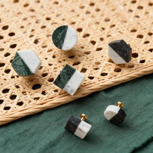 Könnte beinhalten: Eine Sammlung dekorativer Schrankknöpfe in verschiedenen geometrischen Formen. Die Knöpfe weisen eine Kombination aus weißem und dunkelgrünem oder schwarzem Marmor auf. Einige Knöpfe haben einen goldfarbenen Sockel. Die Knöpfe sind auf einer gewebten Oberfläche und einem grünen Stoff angeordnet.