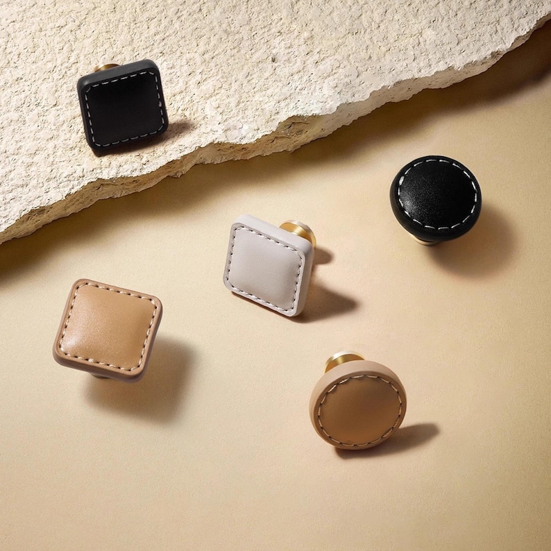 Może przedstawiać: Asortyment sk&oacute;rzanych gałek do szafek w r&oacute;żnych kształtach i kolorach. Gałki mają kwadratowe i okrągłe wzory w kolorze czarnym, beżowym i kremowym, z białymi przeszyciami i złotym osprzętem. Te dekoracyjne gałki są idealne do mebli.