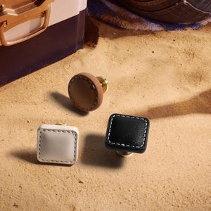 Może przedstawiać: Trzy dekoracyjne gałki do szafek w neutralnych odcieniach. Jedna jest okrągła, ze sk&oacute;ry w kolorze jasnobrązowym z mosiężną podstawą. Pozostałe dwie są kwadratowe, jedna biała i jedna czarna, z białymi przeszyciami i mosiężnymi podstawami. Wszystko na piaszczystej powierzchni.