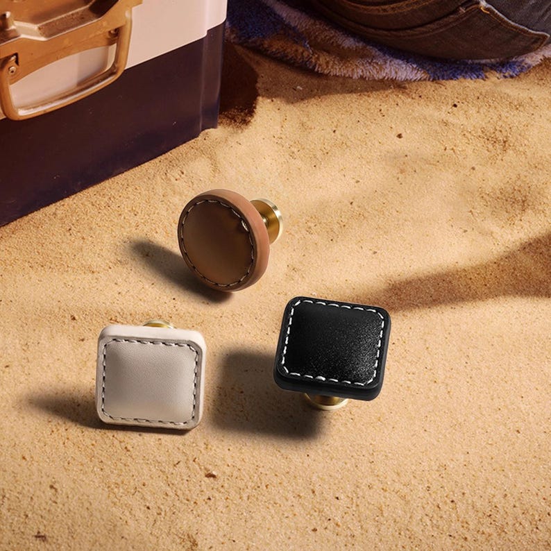 Może przedstawiać: Trzy dekoracyjne gałki do szafek w neutralnych odcieniach. Jedna jest okrągła, ze sk&oacute;ry w kolorze jasnobrązowym z mosiężną podstawą. Pozostałe dwie są kwadratowe, jedna biała i jedna czarna, z białymi przeszyciami i mosiężnymi podstawami. Wszystko na piaszczystej powierzchni.