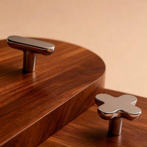May include: Two chrome cabinet knobs, one T-shaped and one cross-shaped, sit on a dark wood surface. The knobs have a shiny, reflective finish. The wood has a rich, warm brown color and a visible grain pattern. The background is a neutral beige.