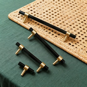 Puede incluir: Cuatro tiradores de armario con un diseño moderno. Los tiradores tienen un cuerpo negro cilíndrico con detalles dorados y tiradores en forma de T. Los tiradores están dispuestos sobre una superficie verde con un fondo de mimbre tejido.