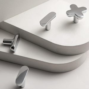 May include: A collection of polished chrome cabinet knobs in various shapes. The knobs include a cross shape and several oblong designs. The hardware is displayed on a white surface with a gray edge, creating a modern aesthetic.