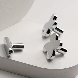 May include: Three shiny silver cufflinks with a clover-like design. The cufflinks are displayed on a white surface, with a soft, diffused background. The design is modern and minimalist, reflecting light.