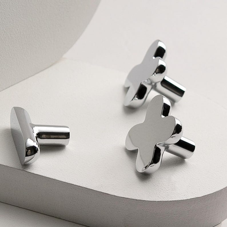 May include: Three shiny silver cufflinks with a clover-like design. The cufflinks are displayed on a white surface, with a soft, diffused background. The design is modern and minimalist, reflecting light.