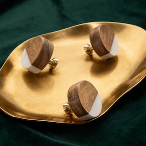 May include: Three cabinet knobs with a two-tone design. The knobs combine light wood and white accents, mounted on brass hardware. They are arranged on a gold-coloured tray against a dark green velvet background.