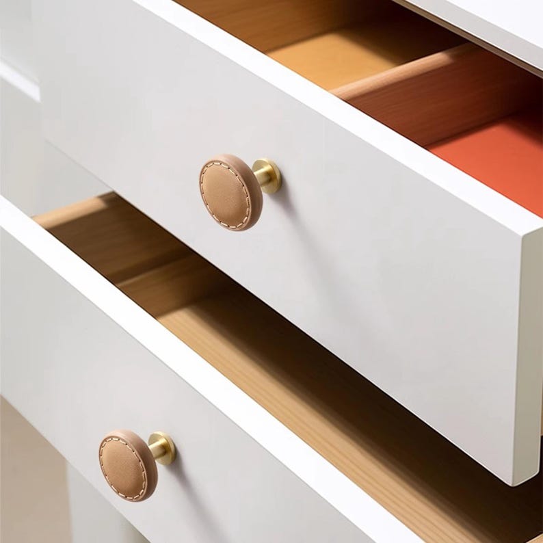 Może przedstawiać: Zbliżenie białej komody z trzema szufladami. Każda szuflada ma okrągłą, brązową sk&oacute;rzaną gałkę z mosiężnym osprzętem. Wnętrza szuflad są w kolorze drewna, z pomarańczowym akcentem.