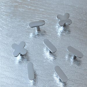 May include: A collection of silver-toned cabinet knobs in various shapes, including rounded rectangles and cross-like designs. The knobs are displayed on a textured, silver surface, showcasing their reflective quality and modern aesthetic.