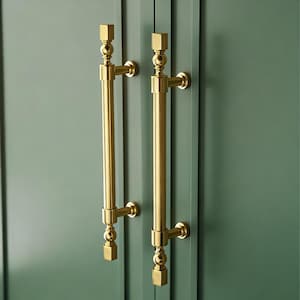 May include: Two gold-colored cabinet pulls with a vertical bar design. Each pull has a square top and bottom, with a decorative sphere and a cylindrical bar in between. The pulls are mounted on a green cabinet door.