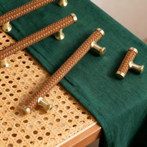 Può includere: Primo piano di diverse maniglie per mobili in pelle marrone con un design intrecciato e ferramenta color oro. Le maniglie sono disposte su un panno verde e una superficie intrecciata marrone chiaro. Le maniglie sono un elemento decorativo per mobili.