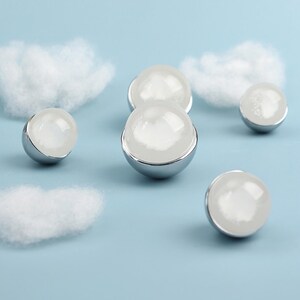 May include: Several spherical, clear glass objects with a white interior and a silver-colored base are arranged on a light blue background with fluffy white cloud shapes. The objects are reflective and appear to be decorative.
