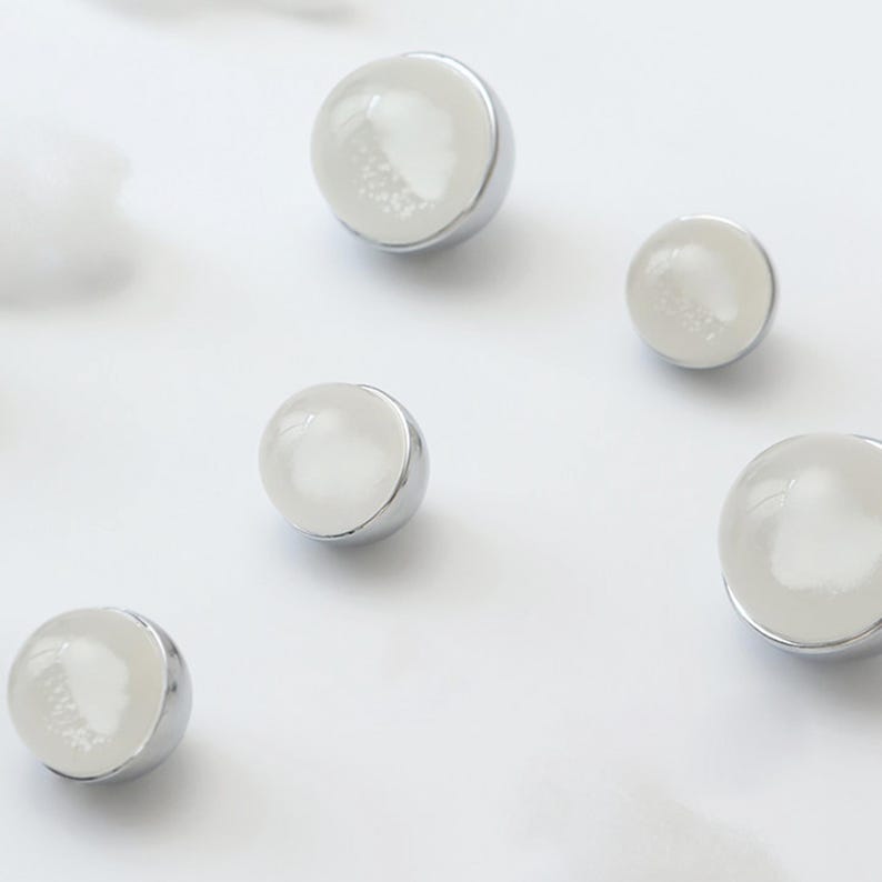 May include: A collection of small, round earrings with a silver-colored base and a white, pearlescent top. The earrings are scattered on a white surface, showcasing their simple, elegant design. These earrings are perfect for everyday wear.
